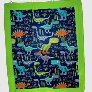May include: A blue fleece baby blanket with a green border. The blanket features a repeating pattern of colorful dinosaurs with the text "I'm Big", "I'm Hungry", "I'm Tall", "I'm Spiky", "I'm Fast", and "Roarrrr".