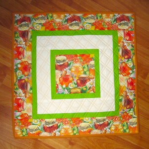 May include: A square quilted placemat with a honey theme. The center features a square of honey jars and flowers. It is surrounded by a white square, a green square, and a border with honey jars and floral patterns. The edges are orange.