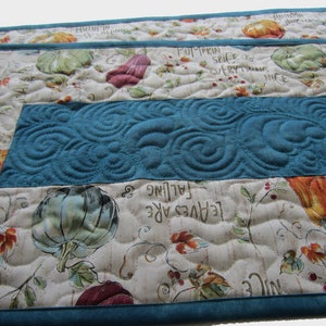 May include: A quilted table runner with a teal blue center panel and a white background with a fall theme. The runner features a repeating pattern of pumpkins, leaves, and the text "Leaves are falling" and "Pumpkin spice is everything nice".