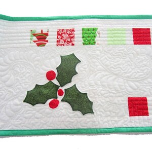 May include: White quilted table runner with a green border. The runner features a green holly leaf design with red berries and a red and green Christmas tree design.