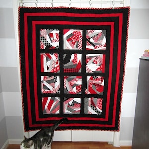May include: A red, black, and white patchwork quilt with a geometric design. The quilt is hanging on a white door with a white rod and black rings.
