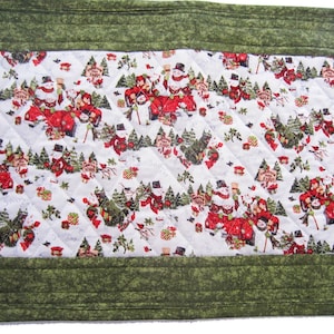 May include: A green quilted table runner with a white background featuring a repeating pattern of snowmen and Christmas trees. The snowmen are wearing red hats and scarves and are pulling a red wagon filled with presents.