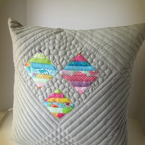 K&ouml;nnte beinhalten: Ein grau gestepptes Kissen mit einem Rautenmuster. In der Mitte befinden sich drei farbenfrohe Rautenformen aus Patchwork-Stoff. Die Patchwork-Rauten haben verschiedene Muster und Farben, darunter Blau, Rosa, Gr&uuml;n und Gelb. Das Kissen ist ein dekoratives Wohnaccessoire.