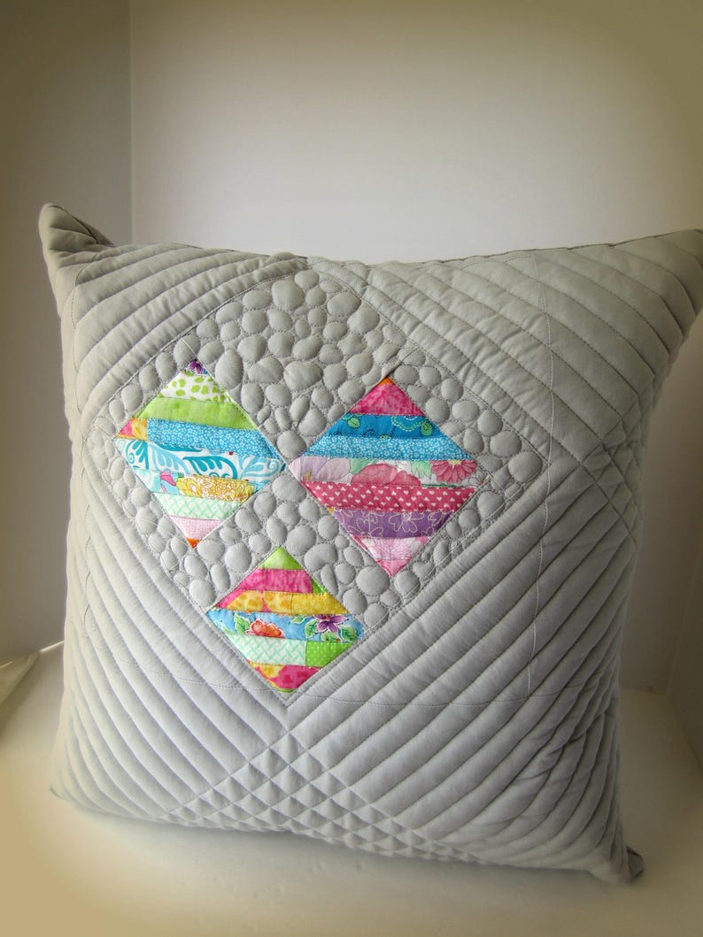 K&ouml;nnte beinhalten: Ein grau gestepptes Kissen mit einem Rautenmuster. In der Mitte befinden sich drei farbenfrohe Rautenformen aus Patchwork-Stoff. Die Patchwork-Rauten haben verschiedene Muster und Farben, darunter Blau, Rosa, Gr&uuml;n und Gelb. Das Kissen ist ein dekoratives Wohnaccessoire.