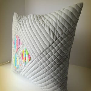 K&ouml;nnte beinhalten: Ein quadratisches, hellgraues, gestepptes Kissen mit einem geometrischen Muster. Das Kissen hat ein Patchwork-Design mit bunten vertikalen Streifen in Rosa, Blau, Gelb und Gr&uuml;n. Die Steppung erzeugt eine strukturierte Oberfl&auml;che.