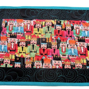 May include: A black quilted table runner with a turquoise border features a colorful pattern of nutcracker figurines. The runner is quilted with a swirling design.