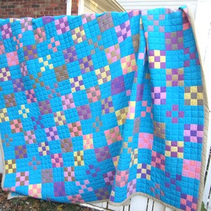 May include: A patchwork quilt with a blue background and squares of pink, purple, yellow, and brown. The quilt is hanging on a white fence.