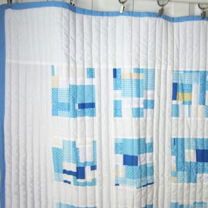 May include: A white quilt with a blue border and a patchwork design of blue and white squares. The quilt is hanging on a white wall.