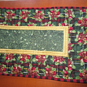 May include: A green and red plaid table runner with a gold trim and a green floral design in the center. The runner has a black and red checkered border.
