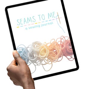 May include: A black tablet shows the text "SEAMS TO ME" above "a sewing journal." Below the text are colourful skeins of thread in shades of blue, white, yellow, orange, and pink. A hand holds the tablet.