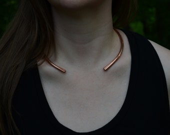 May include: A simple copper choker necklace. The necklace is made of a single piece of copper wire that is bent into a circle. The ends of the wire are left open and are not joined together.