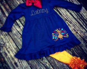 May include: A blue long-sleeved dress with a ruffled hem and a turkey embroidery on the side. The dress is personalized with the name "Lainey".