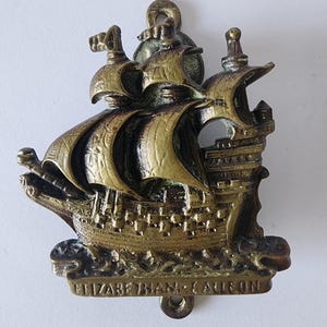 May include: A bronze-coloured ship-shaped wall hanging. The ship features detailed sails, a deck, and a decorative base. The text "ELIZABETHAN - CALLEON" is inscribed on the base. It has a loop at the top for hanging.