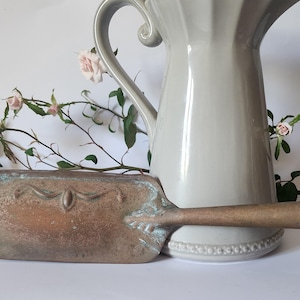 May include: A vintage copper and brass dustpan with a long handle, positioned in front of a grey ceramic jug. The dustpan features an ornate design and a weathered finish. Pink roses and foliage are in the background.