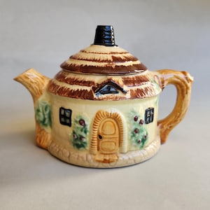 May include: A ceramic teapot shaped like a cottage with a thatched roof, a central arched door, and small windows. The exterior features cream-coloured walls, brown roof detailing, a tree-branch-shaped handle and spout, and green foliage accents.