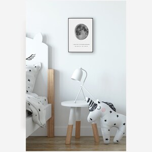 Puede incluir: Una impresi&oacute;n en blanco y negro de una luna llena con el texto "I love you to the moon and back 37&deg; 48' 47" N 127&deg; 12' 37" W" colgada en una pared blanca en una habitaci&oacute;n infantil. Una mesita de noche blanca con base de madera tiene una l&aacute;mpara blanca y un juguete de peluche en forma de unicornio en la parte superior.