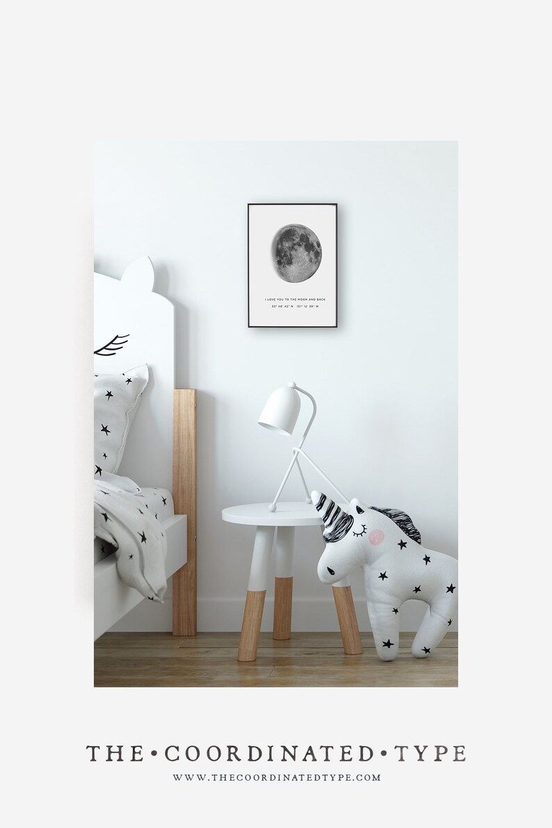 Puede incluir: Una impresi&oacute;n en blanco y negro de una luna llena con el texto "I love you to the moon and back 37&deg; 48' 47" N 127&deg; 12' 37" W" colgada en una pared blanca en una habitaci&oacute;n infantil. Una mesita de noche blanca con base de madera tiene una l&aacute;mpara blanca y un juguete de peluche en forma de unicornio en la parte superior.