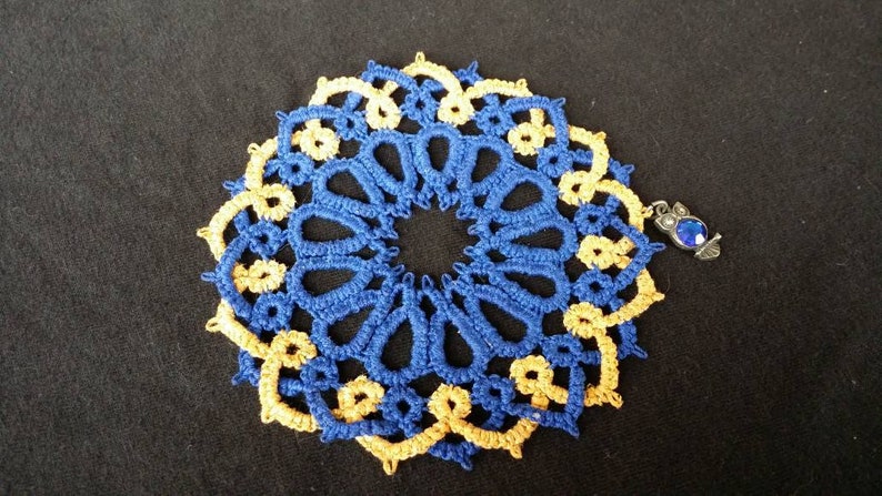 K&ouml;nnte beinhalten: Eine geh&auml;kelte Deckchen in Blau und Gold mit einem kleinen blauen Schmuckanh&auml;nger.