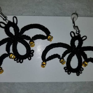 Pode incluir: Um par de brincos de morcego de croch&ecirc; preto com contas douradas. Os brincos est&atilde;o pendurados em ganchos prateados.