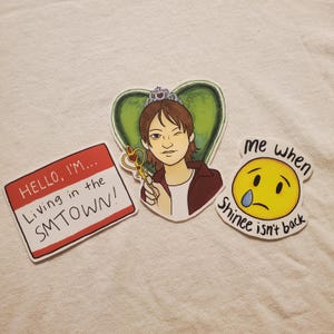 Puede incluir: Tres pegatinas con ilustraciones dibujadas a mano. La primera pegatina es una etiqueta de nombre con el texto "HELLO, I'M... Living in the SMTOWN!" La segunda pegatina es un dibujo animado de una persona con una corona y una llave. La tercera pegatina es una cara sonriente amarilla con una lágrima y el texto "Me when Shinee isn't back".