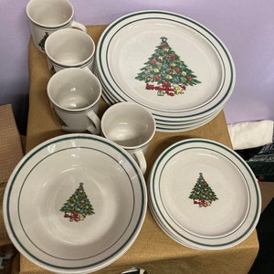 Puede incluir: Un juego de vajilla con temática navideña que incluye platos, cuencos y tazas blancos. Cada pieza tiene un borde verde y un diseño de árbol de Navidad. El juego incluye platos, cuencos y tazas, perfecto para comidas festivas.