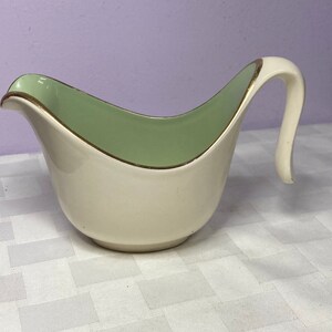 Peut inclure: Un saucier en céramique blanche avec un intérieur vert et un bord doré. La poignée est en forme de bec verseur incurvé.