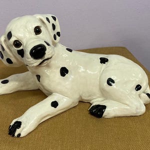 Puede incluir: Una figurita de perro dálmata de cerámica con manchas negras sobre fondo blanco. El perro está acostado con la cabeza girada hacia un lado.
