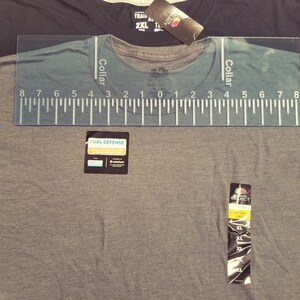 May include: Close-up of three t-shirts, one dark gray, one charcoal, and one black. A clear ruler is placed over the collar of the gray shirt. Tags are attached to the shirts, including "FRUIT OF THE LOOM SELECT" and "DUAL DEFENSE".