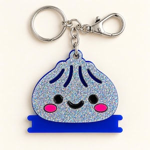 Puede incluir: Un llavero de metal plateado con un dije de dumpling azul brillante con cara sonriente, ojos negros y mejillas de color rosa intenso. El dije está unido a una anilla circular y un mosquetón, con una base rectangular azul sólida debajo.