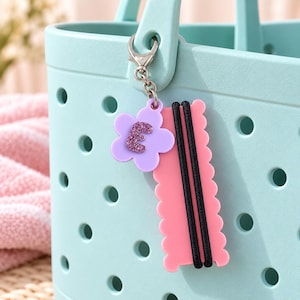 May include: A light blue perforated tote bag featuring a hanging keychain accessory. The keychain includes a lavender flower-shaped charm with a glittery purple letter E and a pink scalloped rectangular tag holding two black elastic hair ties.