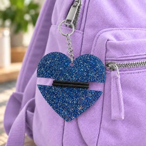May include: A blue glitter heart-shaped keychain attached to a light purple canvas backpack. The heart features a horizontal black elastic band across the center and is connected to the bag by a silver metal split ring and a short chain link.