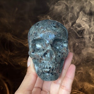 Op de afbeelding: Een grijze en zwarte gevlekte stenen schedel sculptuur. De schedel is gedetailleerd met oogkassen, een neus en een mond.