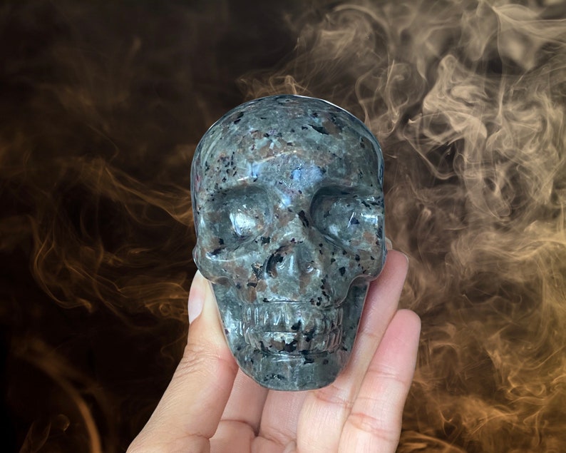 Op de afbeelding: Een grijze en zwarte gevlekte stenen schedel sculptuur. De schedel is gedetailleerd met oogkassen, een neus en een mond.