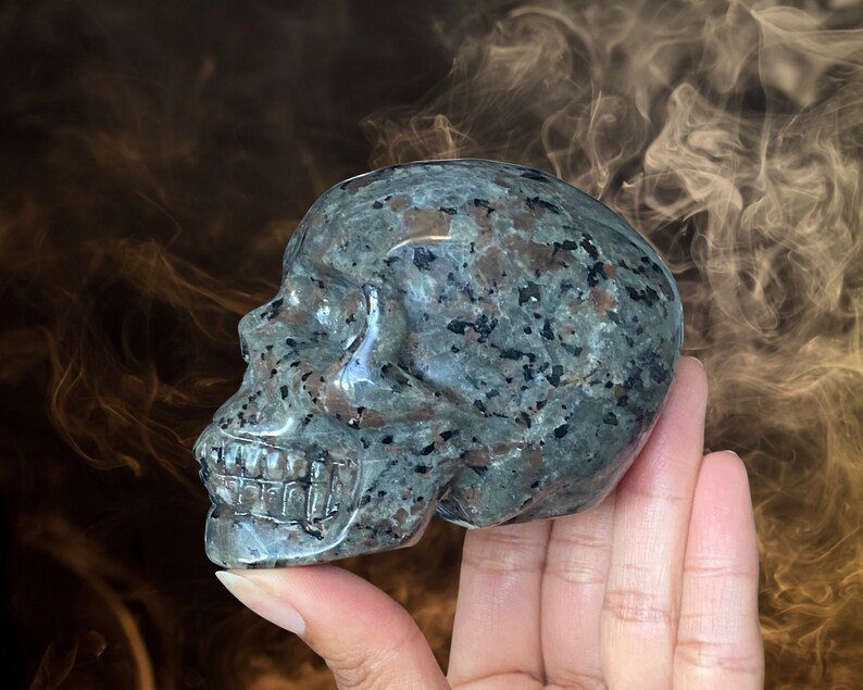 Op de afbeelding: Een donkergrijze en zwart gevlekte stenen schedelsculptuur. De schedel is gedetailleerd gesneden en heeft een gladde, gepolijste afwerking.