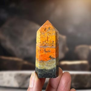 Peut inclure: Un obélisque en pierre précieuse multicolore avec un sommet pointu. La pierre présente des couches de jaune, d'orange, de vert et de noir. L'obélisque est tenu dans une main, mettant en valeur sa surface polie et ses motifs naturels. Cet objet décoratif est un ajout unique.