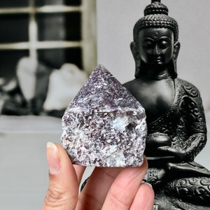 Op de afbeelding: Een paarse en witte kristallen piramide met een ruw, getextureerd oppervlak. Het kristal wordt voor een zwart Boeddha-beeld gehouden. De achtergrond is een wazige zwart-wit afbeelding.