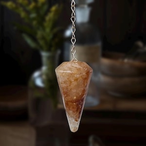 May include: A faceted, teardrop-shaped pendulum made of a translucent, amber-colored crystal. The crystal is suspended from a delicate silver chain. The background is blurred, with hints of greenery and dark wooden surfaces.