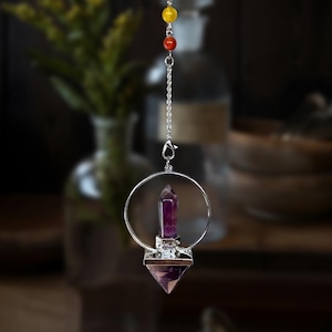 May include: A silver-toned pendulum with a purple crystal point suspended from a chain. The crystal is set within a circular frame and a diamond-shaped base. The chain is adorned with yellow, red, and orange beads. The background is blurred.