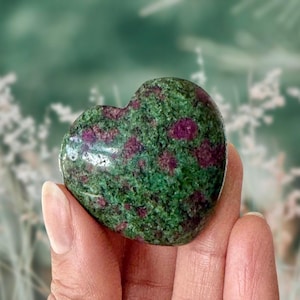 May include: A heart-shaped stone with a green and red speckled pattern. The polished surface of the stone is held in a hand, with a blurred background of green and white foliage.