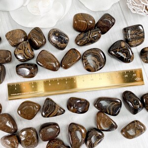 May include: A collection of tumbled brown and black banded jasper stones. The stones are various sizes and shapes, and some have a white stripe running through them. A ruler is included for scale.