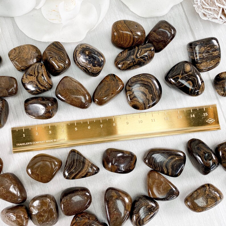 May include: A collection of tumbled brown and black banded jasper stones. The stones are various sizes and shapes, and some have a white stripe running through them. A ruler is included for scale.