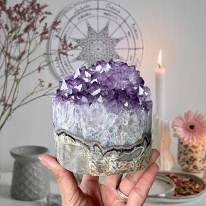 May include: A large, raw amethyst crystal cluster with deep purple points atop a grey and white stone base. The crystal is held in a hand, with a candle and flower in the background. A decorative piece for home decor.