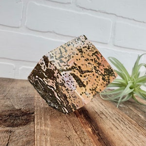 May include: A cube-shaped piece of polished, multi-colored agate with brown, pink, and white swirls and black dendrites. The agate is sitting on a wooden surface.