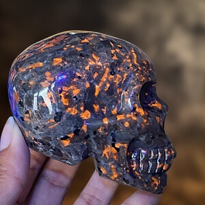 Op de afbeelding: Een zwarte en oranje obsidianen schedel met een gedetailleerde carving van het gezicht en de tanden. De schedel is gepolijst en heeft een glanzende afwerking.