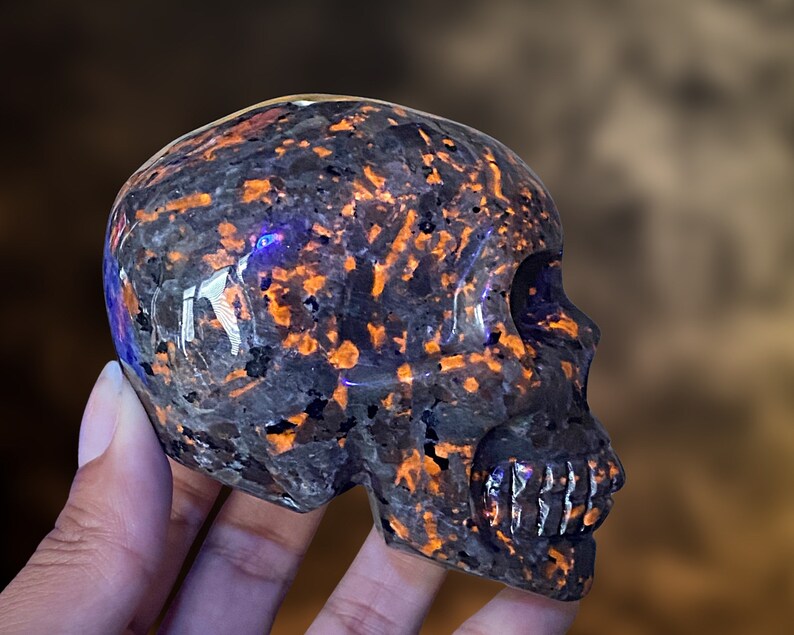 Op de afbeelding: Een zwarte en oranje obsidianen schedel met een gedetailleerde carving van het gezicht en de tanden. De schedel is gepolijst en heeft een glanzende afwerking.