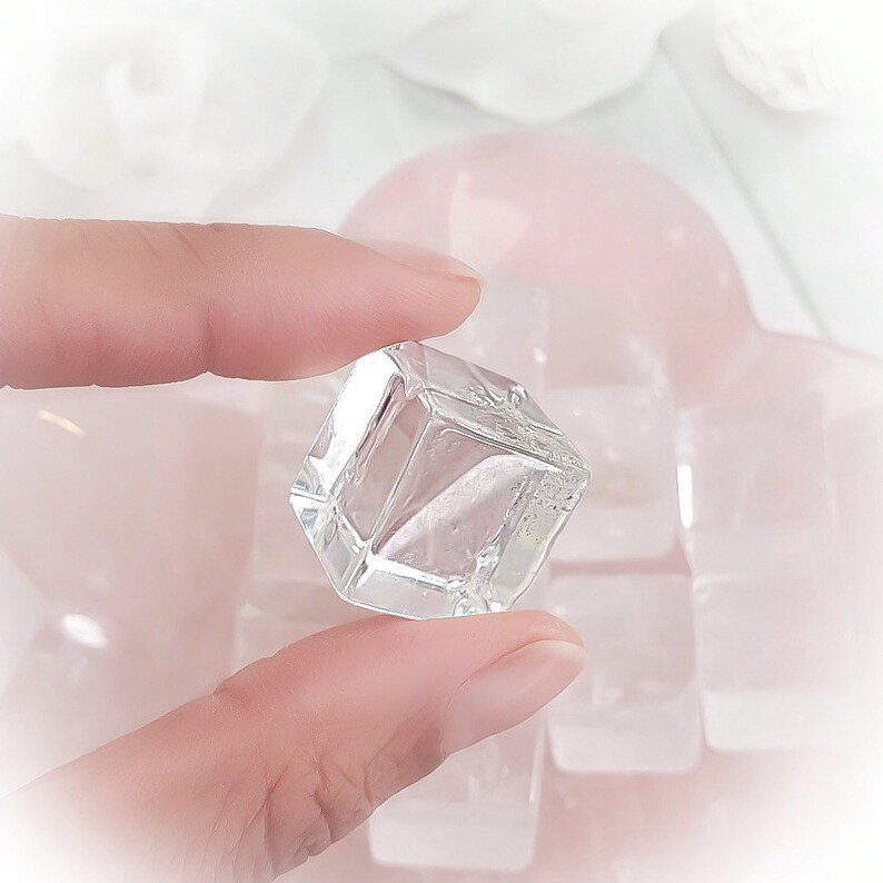 May include: A clear glass ice cube held between two fingers. The ice cube is sitting on top of a pile of other ice cubes.