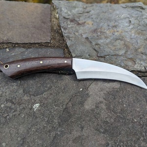 May include: A curved, stainless steel knife with a dark brown wooden handle and brass accents.