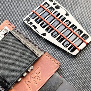 May include: A silver multi-tool with various screwdriver bits, secured by orange rubber bands, rests near a brown leather wallet with the initials "MRF" embossed on it. The wallet has a black elastic band and a metal clasp.