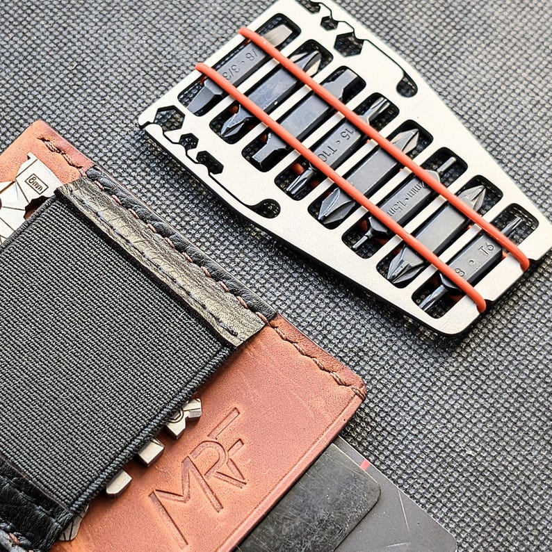 May include: A silver multi-tool with various screwdriver bits, secured by orange rubber bands, rests near a brown leather wallet with the initials "MRF" embossed on it. The wallet has a black elastic band and a metal clasp.