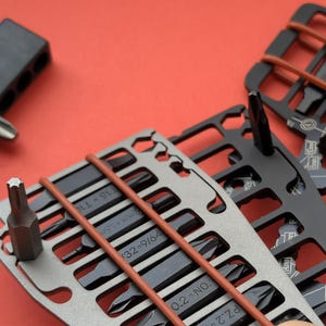 May include: Close-up of a set of multi-tool cards with various screwdriver bits. The cards are silver and black, held together with brown rubber bands, and set against a red background. The bits are black and arranged in rows.